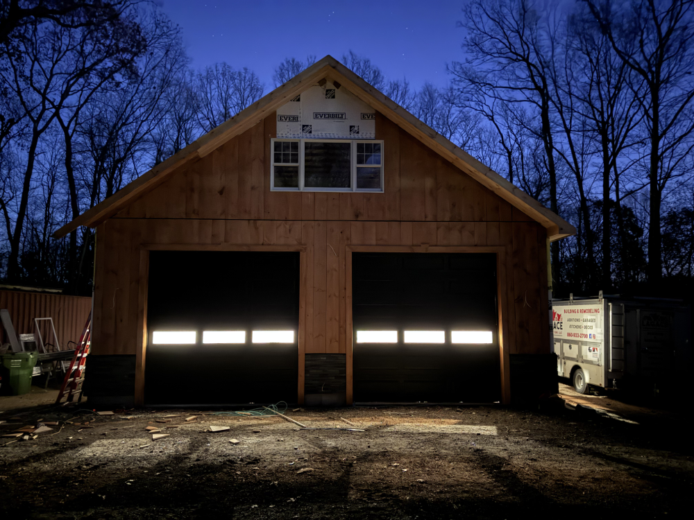 A newly constructed wooden garage at dusk, featuring illuminated garage doors and surrounding trees silhouetted against a dark blue sky.