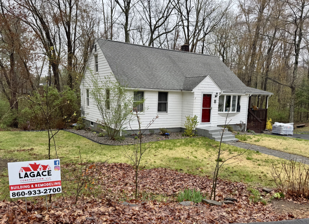 Lagace Remodelling LLC A charming white house with a gray roof, surrounded by trees, featuring a sign for L'Agace Construction, advertising building and remodeling services.