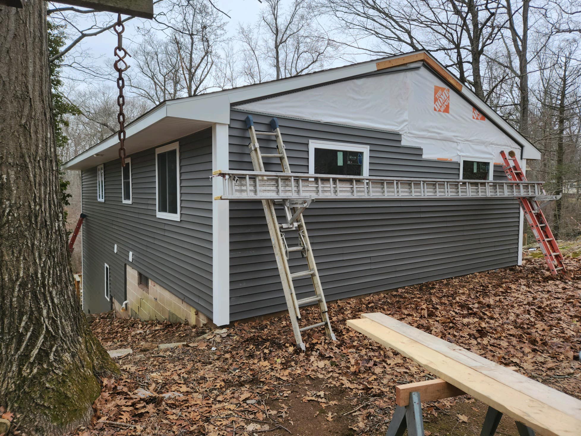 Sloped house getting new siding in andover connecticut House on a sloped lot undergoing exterior renovation with new dark gray vinyl siding being installed. The upper portion is still wrapped in Home Depot-branded house wrap. Several ladders and construction tools are positioned around the site, with fallen leaves covering the ground. Located in Andover, Connecticut.